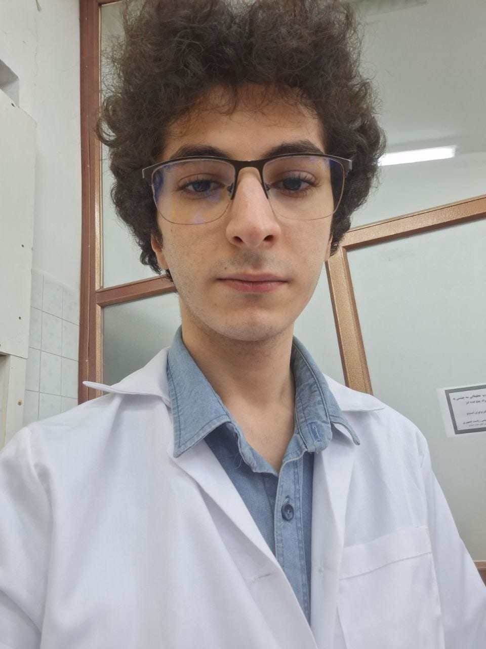 Afshin Ghorbani in a white lab coat over a denim shirt, head and shoulders, standing in a laboratory corridor.